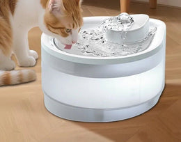 Ginger and white cat drinking from a round white running-water cat fountain with a bubbling centre spout, on a wooden floor