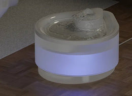 White cat water fountain glowing with a soft blue-white night light on a dark wooden floor beside a rug