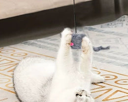 White cat lying on its back on a patterned rug, holding a grey furry mouse that hangs on a cord