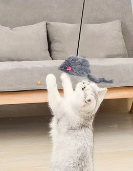Pale grey cat standing on its back legs to reach a grey furry mouse hanging on a cord in front of a sofa