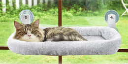 Tabby cat lying in a grey fluffy window bed held to the glass by two suction-cup brackets, with a garden outside