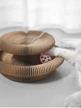 Cardboard accordion scratcher opened into a two-tier ring with a red bell ball, and a white cat paw reaching in