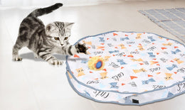 Tabby kitten pouncing at a ball in a fluffy yellow cover beside the cloth play pouch on a pale floor