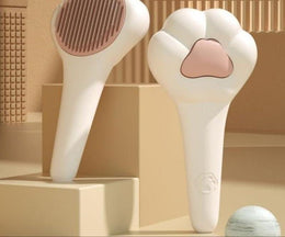 Front and back of the white paw-shaped cat brush on beige blocks: its round pin head, and its back with a pink paw pad