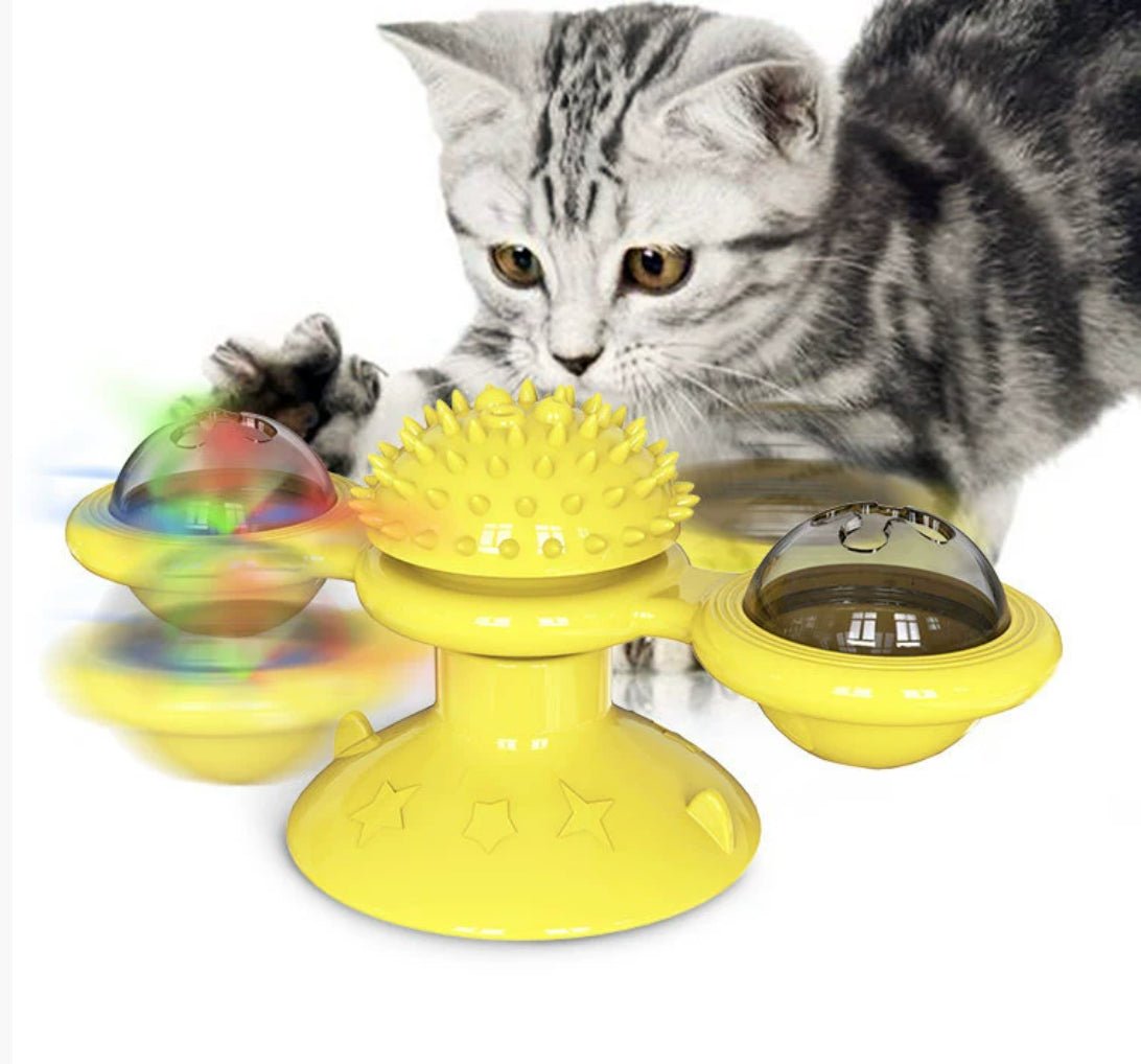 Silver tabby kitten pawing at a yellow windmill toy on a suction-cup base, with a spiky centre and a clear dome at each end of the arm, one lit up in several colours